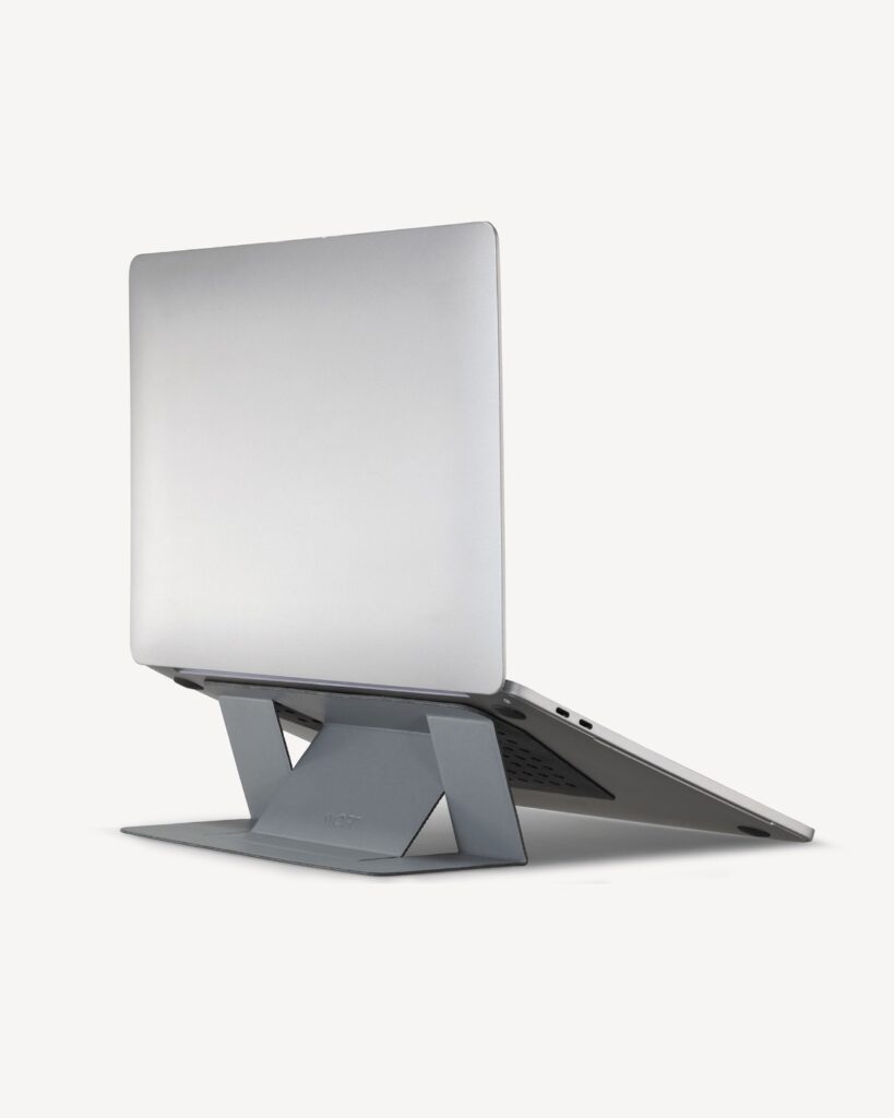Complete silver laptop supported by the grey MOFT Adhesive Laptop Stand with the folding structure fully visible